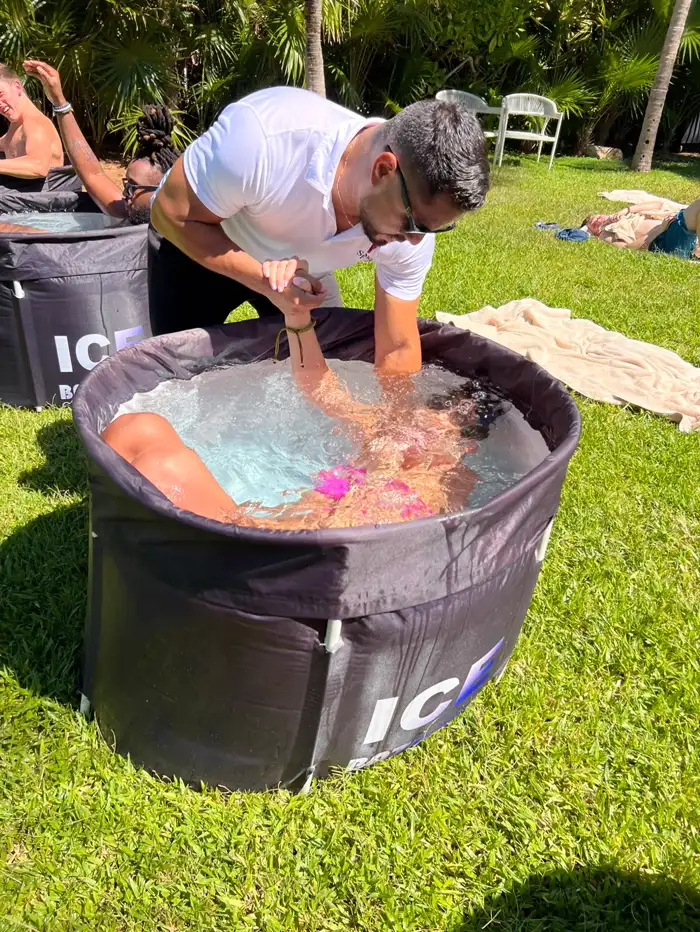 El guía, de pie junto a la tina, le sostiene las manos a una huésped sumergida en el agua con hielo