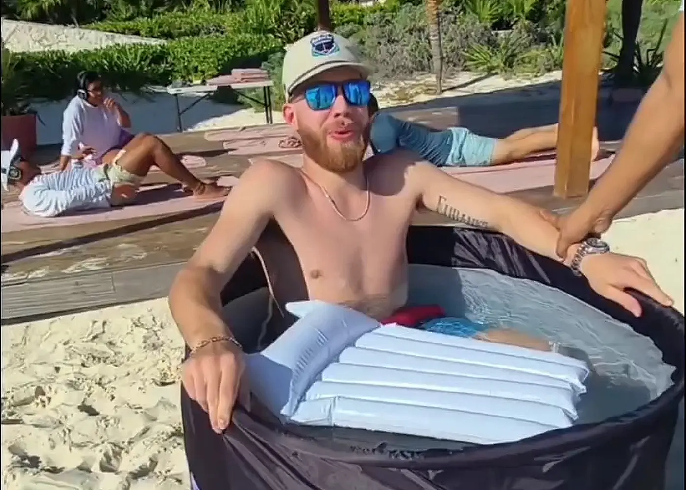 A guest sitting in an ice bath tub set up on the sand under a palapa, with the resort behind
