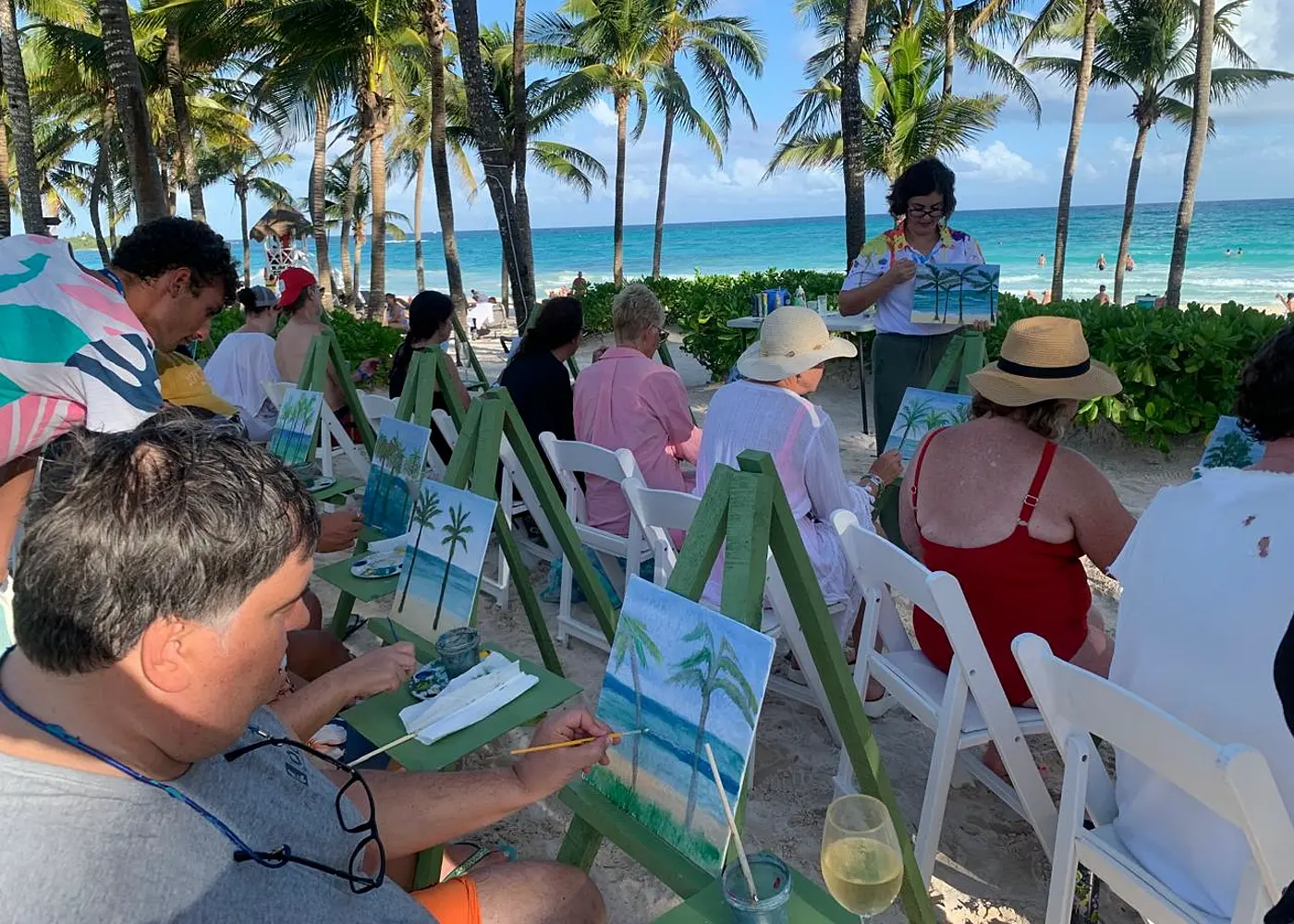 A row of green easels set up on the sand facing the sea in Tulum, guests painting palm trees while a Splat artist stands guiding the class